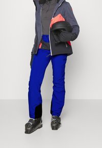 Personne portant un pantalon de ski bleu, des bottes de ski noires, des gants noirs, une veste grise et rouge, tenant un casque de ski noir.
