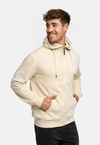 Felpa a zip color beige chiaro realizzata in tessuto morbido, dotata di cappuccio con coulisse, due tasche frontali e polsini a costine. Design elegante e casual.