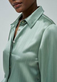 Camisa de satén verde claro con rayas plateadas verticales, que presenta un cuello con solapa, botones en la parte delantera y una textura suave y brillante.