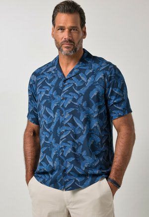 Homme aux cheveux courts et foncés et à la barbe porte une chemise bleue à manches courtes boutonnée avec des motifs de feuilles et un pantalon beige, debout les mains dans les poches.