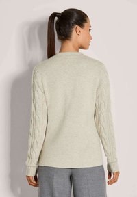 Frau mit einem hohen Pferdeschwanz, die einen hellbeigen Pullover mit Kabelstrickärmeln und grauen Hosen trägt, steht vor einer schlichten Wand.