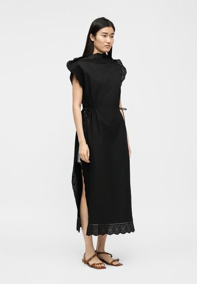Γυναίκα που φοράει μαύρο midi φόρεμα χωρίς μανίκια με δαντέλα eyelet στο τελείωμα, ψηλό λαιμό, πλαϊνές σχισμές και καφέ επίπεδα σανδάλια, στέκεται μπροστά σε απλό φόντο.