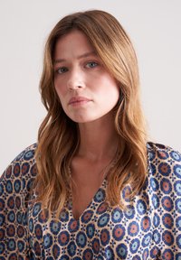 Blumenmusterkleid mit V-Ausschnitt, fließenden Ärmeln und einer Mischung aus Blau, Orange und Beige auf einem glatten Stoff.