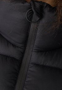 Gilet matelassé noir avec un col haut, un quilting vertical et une fermeture éclair avec une tirette ronde. Matériau en nylon texturé.
