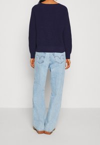 Kvinna klädd i en mörk marinblå tröja och ljusblå högmidjade vida jeans som står vänd bortåt mot en enkel bakgrund.