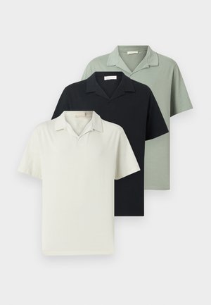 Muškarac obučen u crnu košulju kratkih rukava s ovratnikom, bež hlače i crne mokasine stoji sa rukama iza leđa na bijeloj pozadini.