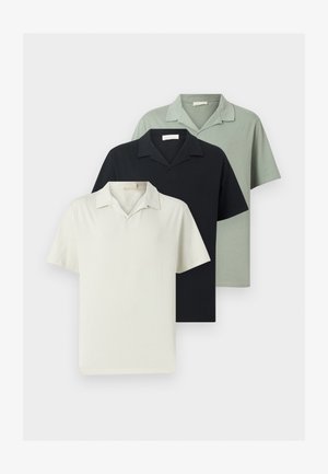 Férfi fekete rövid ujjú galléros ingben, bézs nadrágban és fekete loaferekben áll, kezei a hát mögött, fehér háttér előtt.