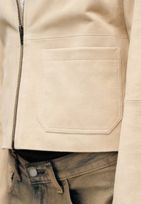 Μπεζ σατέν μπουφάν με μεγάλο μπροστινό τσέπη και μαύρο φερμουάρ, φορεμένο πάνω από ξεθωριαμένα μπλε τζιν με κλείσιμο με κουμπί.