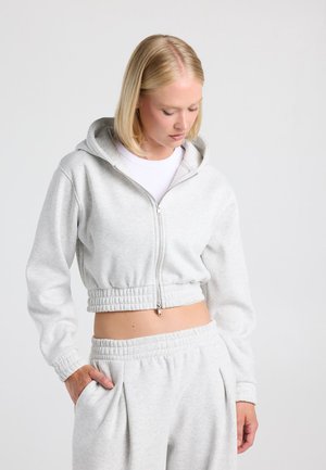Femme aux cheveux blonds portant un sweat à capuche court gris clair avec fermeture éclair et un pantalon de survêtement assorti, les mains dans les poches sur fond uni.