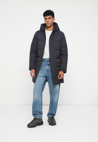 Ung mand iført en sort hættepufjakke, hvid t-shirt, løse blå jeans og sorte sneakers, der står foran en hvid baggrund.