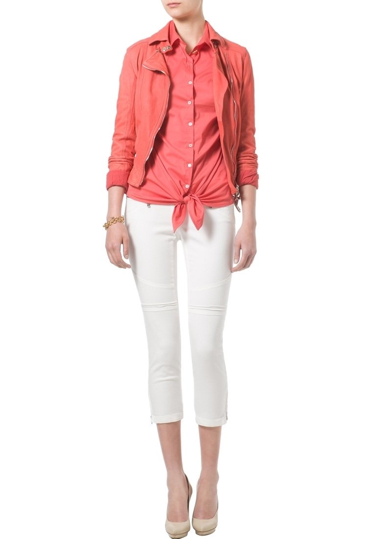 Chaqueta de imitación de cuero color coral sobre una blusa coral anudada, combinada con pantalones capri blancos y tacones nude. Textura suave y diseño ajustado.