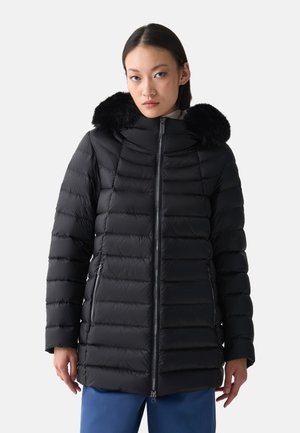 Giovane donna che indossa un cappotto imbottito nero trapuntato con cappuccio bordato di pelliccia e pantaloni blu, in piedi davanti a uno sfondo chiaro e uniforme.