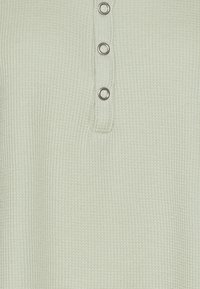 Ljust grön waffle-struktur topp med en knappad henley-krage med fem silverfärgade knappar. Mjuk, texturerad tyg med ribbat detalj.