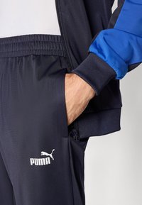 Moški nosi temno modre Puma trenirke in jakno, z roko v žepu, kar kaže na elastični pas in manšeto jakne v modri barvi.