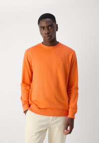 Oranssi huppari pyöreällä kaula-aukolle ja ribbireunuksilla. Rinnassa on huomaamaton logotyyppi. Pehmeä kangas, rento malli ja yksivärinen.