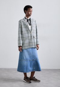 Blazer à carreaux avec un seul bouton, porté sur un chemisier blanc et une jupe en jean. Chaussures marron texturées complètent la tenue.