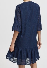 Sininen mekko ohuesta materiaalista, jossa on teksturoitu lehtikuva, kolmen neljäsosan hihaiset röyhelöt sekä leveä helma.