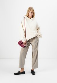 Vrouw in een crèmewitte oversized hoodie, beige wijde pantalons, zwarte loafers en met een kleine bordeauxrode schoudertas, staande tegen een witte achtergrond.