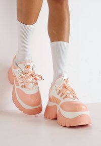 Tömeges sneaker, rózsaszín és fehér patent bőrrel, vastag talppal és bordázott díszítésekkel. Fűzők és egy rajzfilmszerű részlet a nyelvén.