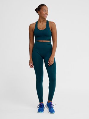 Kvinde smilende, iført teal sports-bh og leggings, stående barfodet på hvid baggrund.