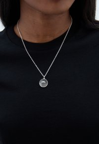 Sudraba kaklarota ar apaļu piekariņu, uz kura ir saules stariņu dizains; nēsāta virs melna T-krekla, izceļot tās metālisko tekstūru.