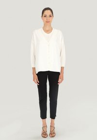 Kremowy kardigan zapinany na guziki z długimi rękawami na jasnej bluzce, w połączeniu z dopasowanymi czarnymi spodniami i sandałami na paski, na neutralnym tle.