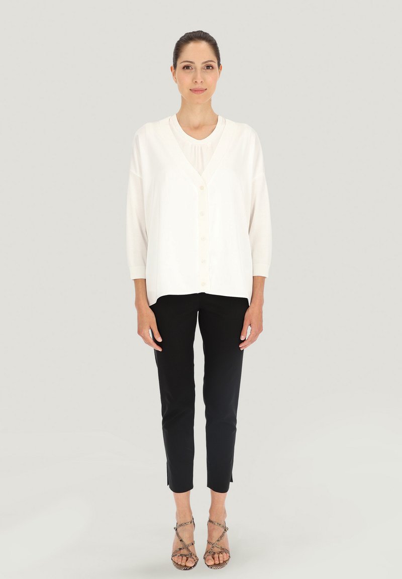 Kremowy kardigan zapinany na guziki z długimi rękawami na jasnej bluzce, w połączeniu z dopasowanymi czarnymi spodniami i sandałami na paski, na neutralnym tle.