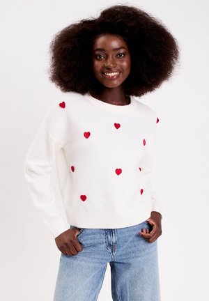 Eine lächelnde Frau mit großen lockigen Haaren, die einen weißen Pullover mit kleinen roten Herzen und hellblauen Jeans trägt, mit den Händen in den Taschen.