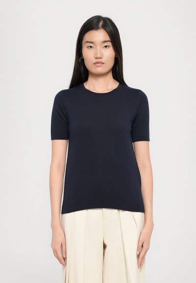 Pull en maille bleu marine à manches courtes avec un col rond et un ourlet côtelé, porté avec un pantalon plissé crème. Texture lisse, design ajusté.