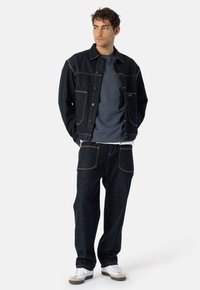 Moški stoji v temnem kavbojskem jopiču in kavbojkah, modri majici ter belih supergah, in gleda vstran na belem ozadju.