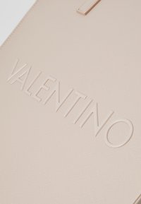 Beige läder tote med präglad "VALENTINO"-logotyp, som har en texturerad yta och minimalistisk design. Ingen anmärkningsvärd hårdvara eller detaljer.