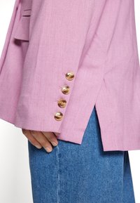 Lys rosa blazer med teksturert stoff, med fire gullknapper langs ermet og en sidesplit for ekstra detalj.