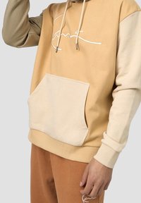 Beige och tan huvtröja med en framficka, dragsko i huva och broderad logotyp. Gjord av mjukt tyg med kontrasterande ärmar.