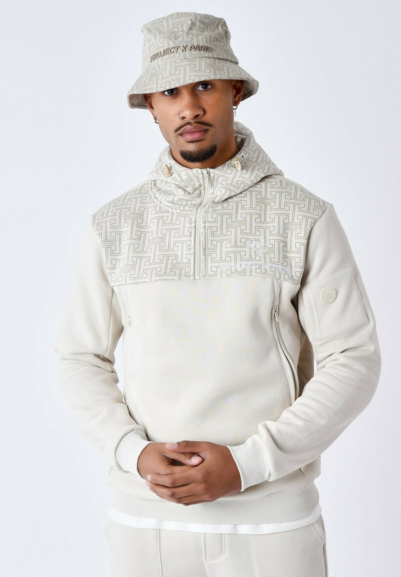 Pullover crème avec une capuche à motif texturé, fermeture éclair à l'avant et poche latérale. Associé à un chapeau de seau assorti, avec un logo discret.