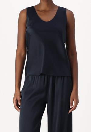 Frau trägt ein ärmelloses marineblaues Oberteil und passende weite Hose vor einem einfarbigen Hintergrund.