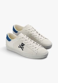 Zapatillas de ante beige con diseño de calavera y tibias en negro, parte delantera con cordones y acento azul en el talón. Suela texturizada en gris.