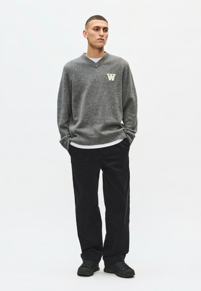Jeune homme debout, les mains dans les poches, portant un pull gris à col en V avec un logo W, pantalon noir et chaussures noires sur fond blanc.