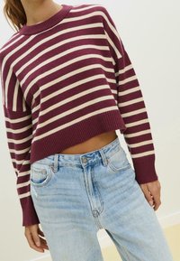Personne portant un pull court rayé bordeaux et blanc associé à un jean taille haute bleu clair sur un fond uni.