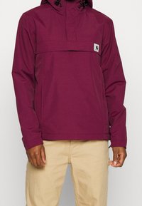 Coupe-vent bordeaux avec une poche avant, une capuche ajustable et un cordon de serrage, fabriqué en matériau lisse et léger. Manches longues et coupe décontractée.