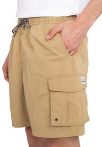 Béžové cargo šortky s elastickým pásom a šnúrkou, obsahujúce veľkú bočnú kapsu a logo nášivku na nohe.