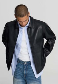 Giovane uomo con i capelli corti che indossa una giacca di pelle nera sopra una camicia a bottoni azzurra chiara e una t-shirt bianca, abbinata a jeans blu, con lo sguardo rivolto verso il basso.
