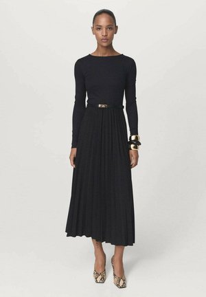 Donna in piedi che indossa un top nero a maniche lunghe con una gonna midi plissettata abbinata, bracciali larghi in oro e nero, e tacchi in pelle con motivo a serpente.