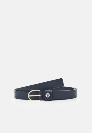 Ceinture en cuir bleu marine avec une boucle en métal doré et un petit emblème rond doré sur la boucle, sur un fond uni.