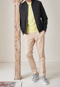 Svart bomberjacka över en gul sweatshirt, i kombination med beige byxor och grå sneakers. Släta material, jacka med dragkedja, avslappnad passform.