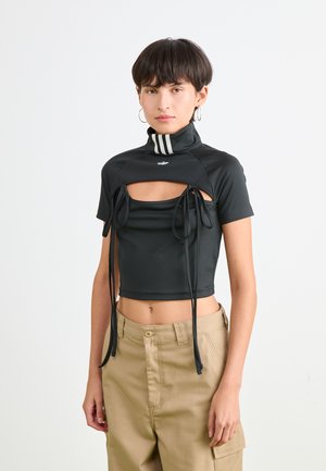 Femme aux cheveux courts et foncés, portant un haut court noir avec découpes et liens, un pantalon beige et des boucles d'oreilles créoles devant un fond blanc uni.