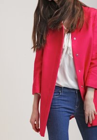 Vaaleanpunainen takki, jossa on tekstuuri, yksittäinen nappisulkiminen ja leveät hihat, päällä valkoinen paita ja siniset farkut.