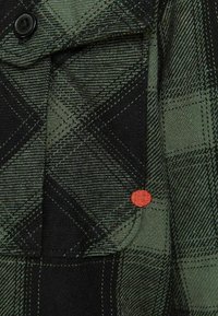 Tessuto in flanella a quadri verde e nero con tasca con bottoni, caratterizzato da un dettaglio a pois arancione. Tessitura texturizzata e forma strutturata.