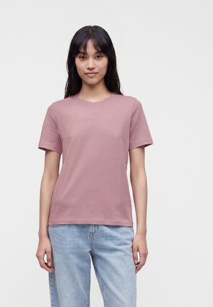 Jeune femme aux longs cheveux noirs portant un t-shirt mauve uni à manches courtes et un jean bleu clair, debout devant un fond blanc.