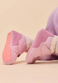 Baby, der weiche rosa und lila Schuhe mit strukturierten Sohlen und cremefarbenen, flauschigen Socken trägt, liegt auf einer beigen Oberfläche.