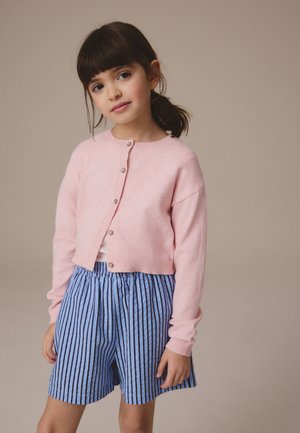 Junges Mädchen mit dunklen Haaren zu einem Pferdeschwanz gebunden, trägt eine hellrosa Knopfleisten-Strickjacke und blaue gestreifte Shorts vor einem einfarbigen beigen Hintergrund.